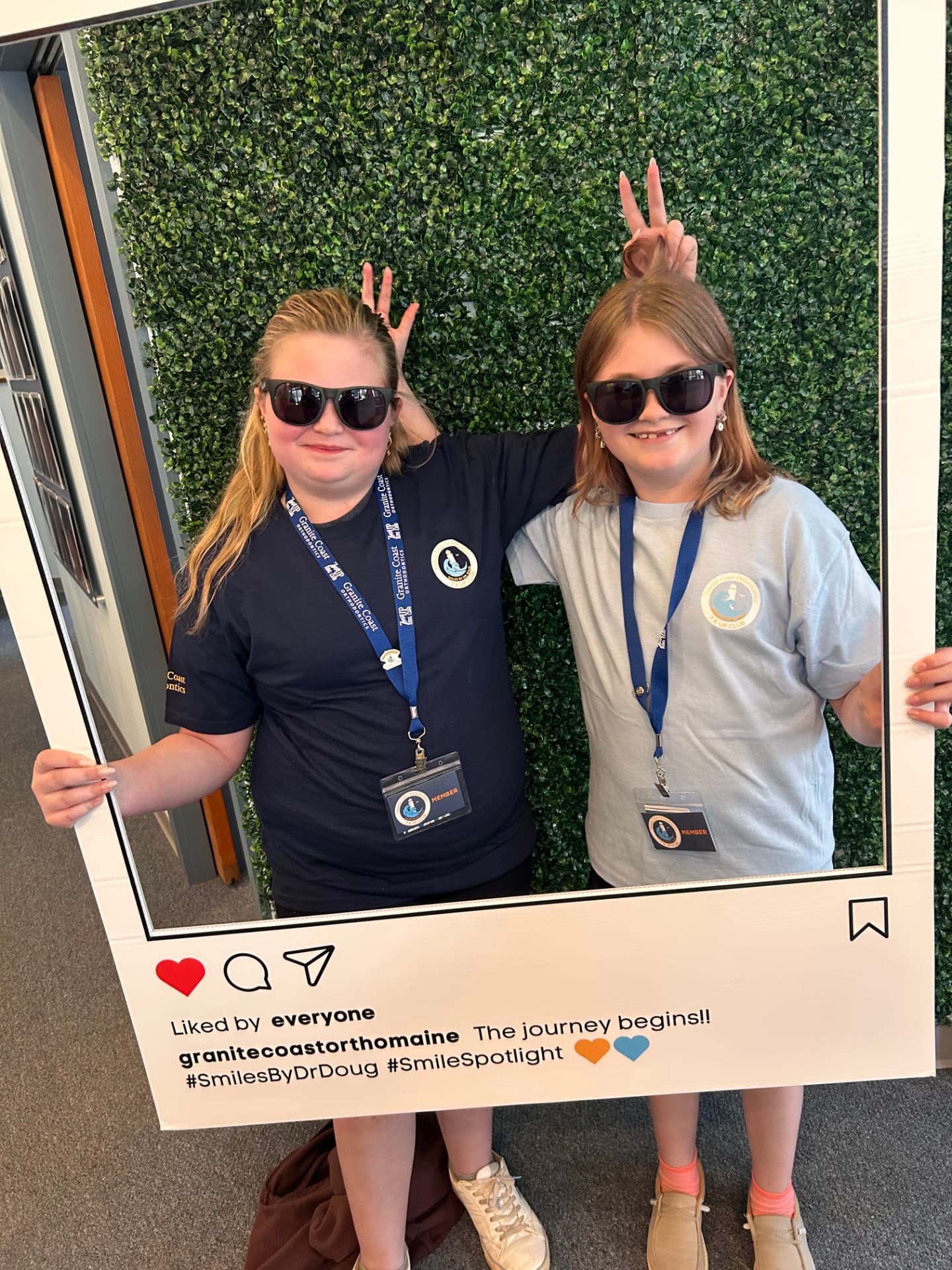 Two 7 & Up Club members in club t-shirts and member badges posing with the Granite Coast Orthodontics photo frame